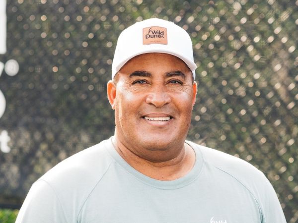 A person wearing a white "Wild Dunes" cap and light gray shirt smiles against a blurred outdoor background.