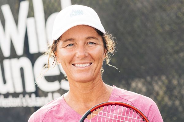 A person in a pink shirt and white cap holds a tennis racket, smiling, with a blurred outdoor background and "Wild Dunes" in text.