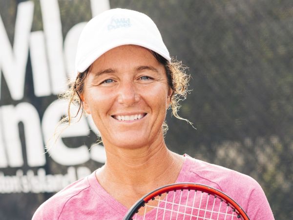 A person in a pink shirt and white cap holds a tennis racket, smiling, with a blurred outdoor background and "Wild Dunes" in text.