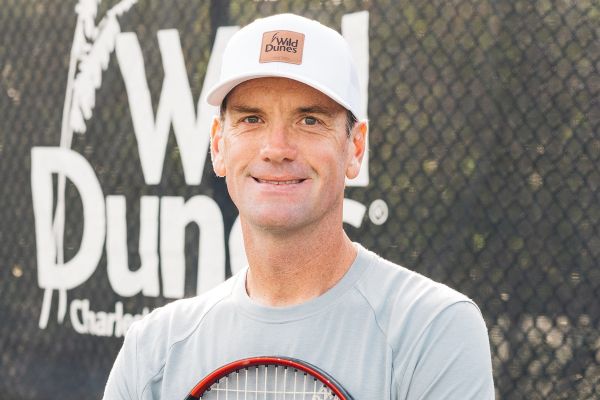 A person in a cap and gray shirt holds a tennis racket, standing in front of a "Wild Dunes" sign on a tennis court background.