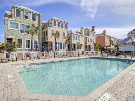 A pool area with lounge chairs is surrounded by colorful multi-story houses and palm trees, under a partly cloudy sky.
