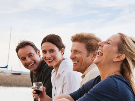 Four people are outdoors near a body of water, smiling and enjoying the moment, with a sailboat visible in the background.