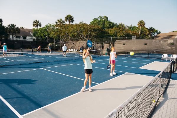Several people are playing pickleball on outdoor courts with blue surfaces, surrounded by trees and buildings in the background.