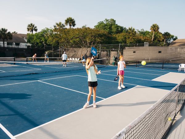 Several people are playing pickleball on outdoor courts with blue surfaces, surrounded by trees and buildings in the background.