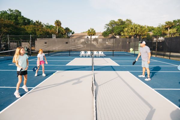 People are playing pickleball on a blue and gray court under the clear sky, surrounded by trees.