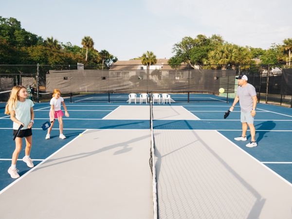 People are playing pickleball on a blue and gray court under the clear sky, surrounded by trees.