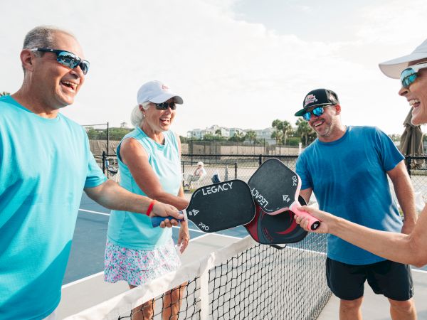 Four people are enjoying pickleball, laughing and touching paddles over a net on a sunny day.