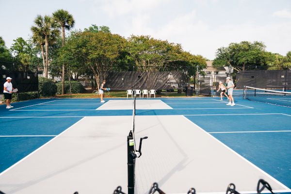 People playing a doubles match on a blue and gray pickleball court, surrounded by trees and a chain-link fence.