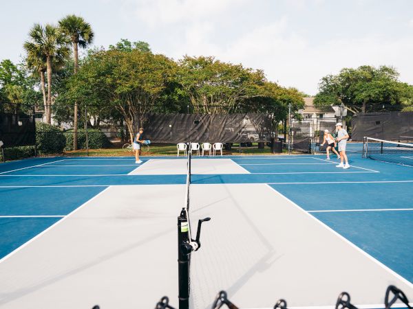 People playing a doubles match on a blue and gray pickleball court, surrounded by trees and a chain-link fence.