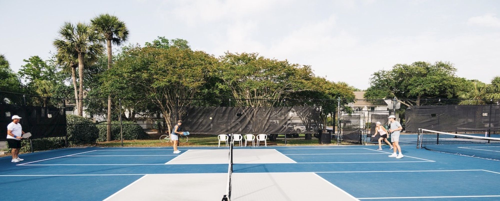 A doubles tennis match is taking place on an outdoor court surrounded by trees, with people actively playing and watching.
