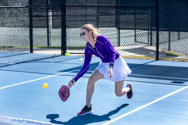 A woman is playing pickleball on a blue court, wearing a purple jacket and white skirt, hitting a yellow ball with a paddle.