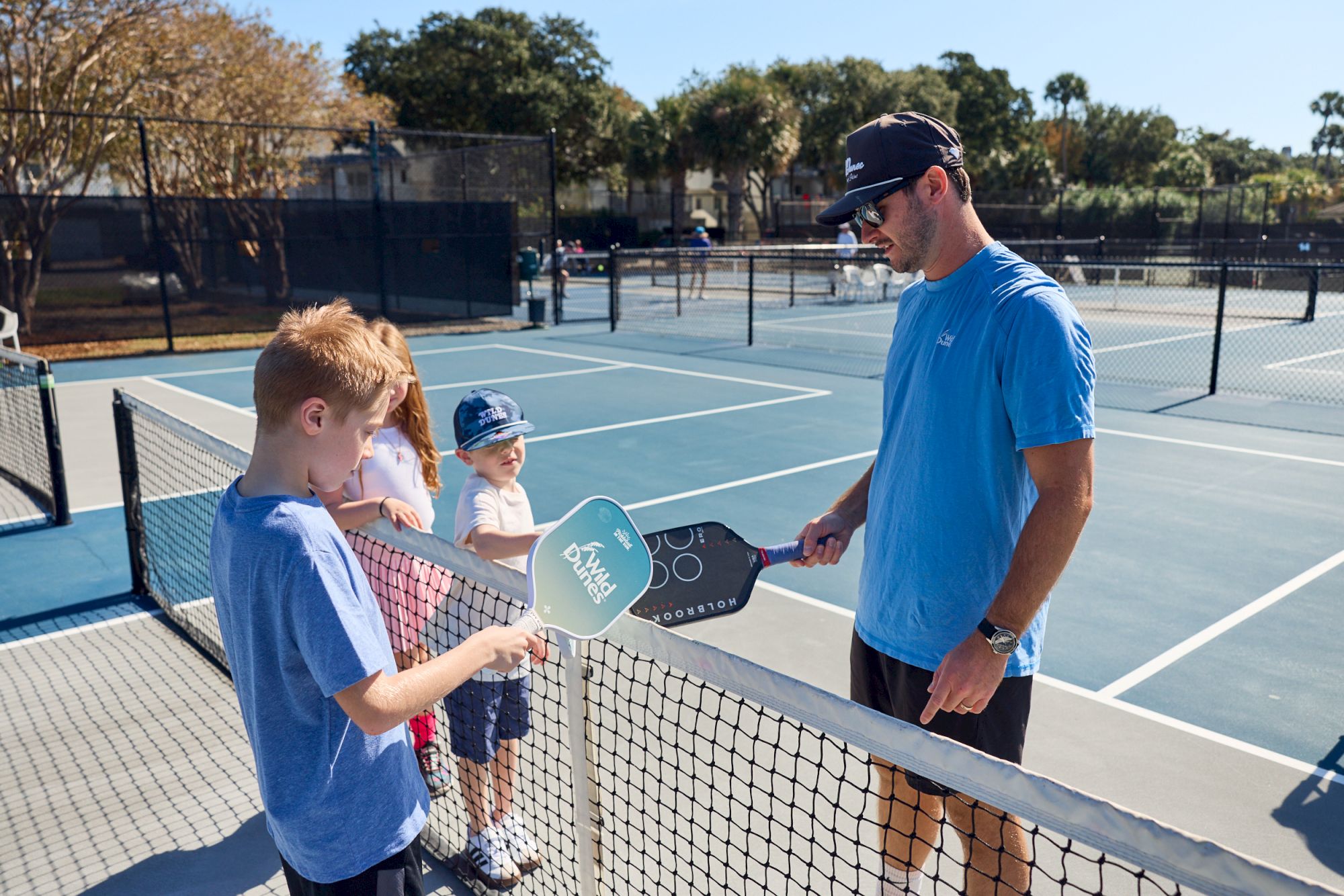Two kids learn paddle‑tennis from a coach at a sunny outdoor court, holding paddles and listening near the net.