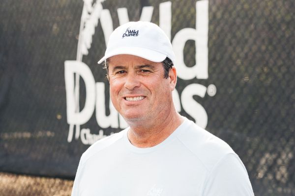 A person wearing a white cap and shirt stands in front of a sign that reads "Wild Dunes" with a tennis court fence in the background.