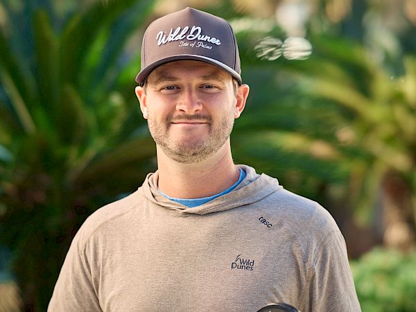 A man wearing a brown hoodie and a cap holds a pickleball paddle and ball outdoors, smiling near green plants.