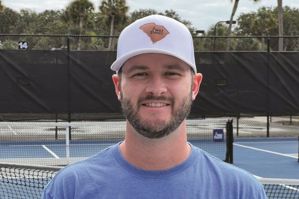 A person wearing a white cap and blue shirt stands in front of a tennis court with nets and a black fence in the background.