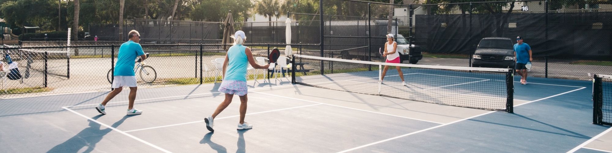 A group of people are playing pickleball on an outdoor court surrounded by a fence and trees in the background.