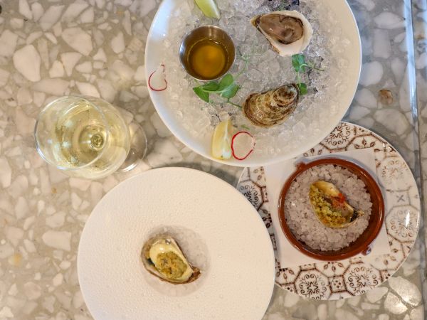 The image shows a table setting with oysters on ice, a glass of white wine, and garnished plates on a patterned surface.