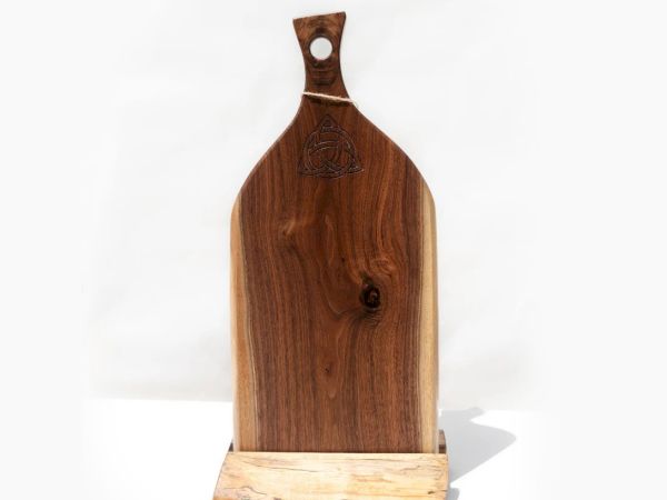 The image shows a wooden cutting board with a handle, featuring a hole for hanging. It's standing upright on a simple wooden base.