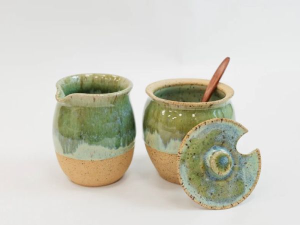 The image shows two ceramic jars in green and brown tones; one has a lid and spoon, possibly handmade pottery.