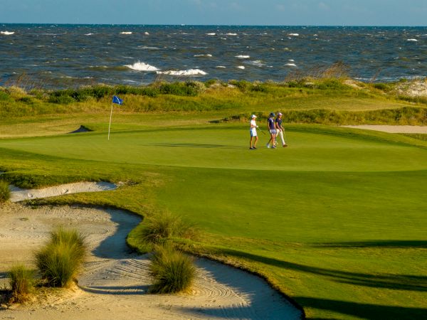 A coastal golf course with people on the green, surrounded by sand traps and waves in the background, under a clear sky.