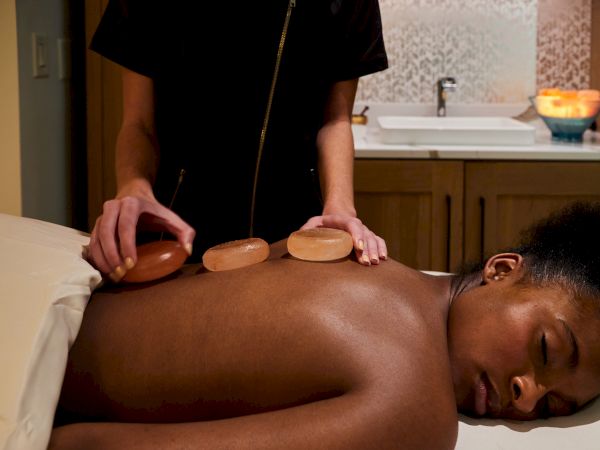A person is receiving a hot stone massage in a spa setting, with stones placed on their back by a massage therapist.