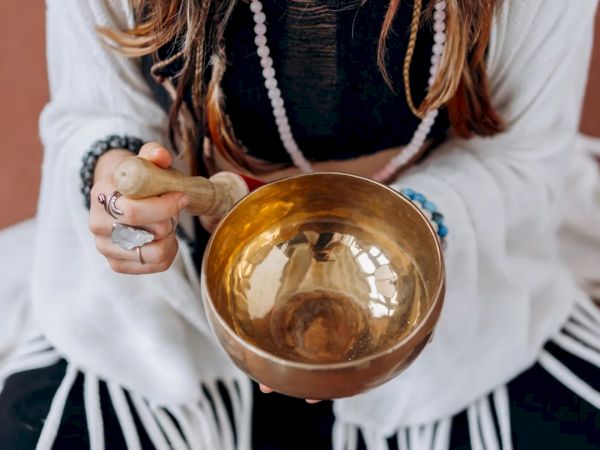 A person holding a singing bowl with a mallet, wearing a white fringed shawl and beaded bracelets.