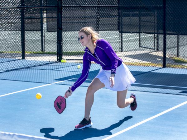 A person in sports attire plays pickleball, hitting a ball with a paddle on an outdoor court, surrounded by a chain-link fence.
