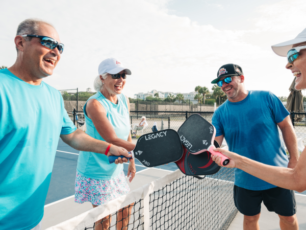Four people smiling and holding pickleball paddles over a net, all wearing sports attire and sunglasses outdoors, enjoying a sunny day.