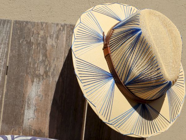 A straw hat with blue and white patterns is placed on a wooden surface next to a cushion with a blue abstract design.