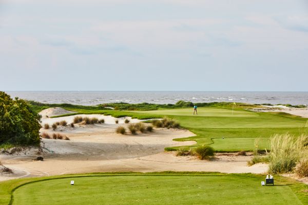 A seaside golf course with sand dunes, grass, and a golfer near the hole, with the ocean in the background.