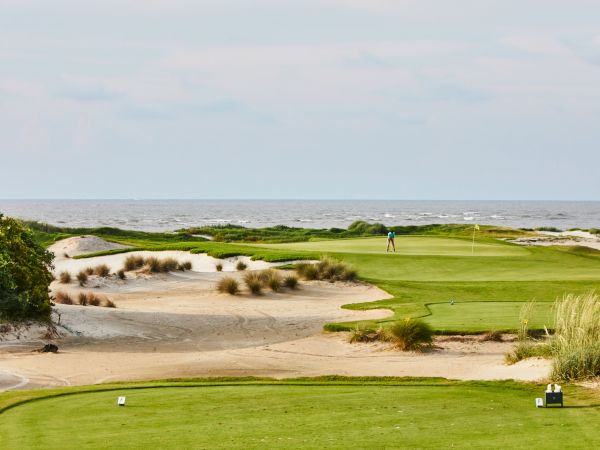 A seaside golf course with sand dunes, grass, and a golfer near the hole, with the ocean in the background.