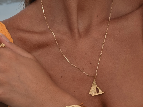 A person wearing a gold necklace with a sailboat pendant, a gold bracelet, and a ring with a small star design, in a relaxed pose.