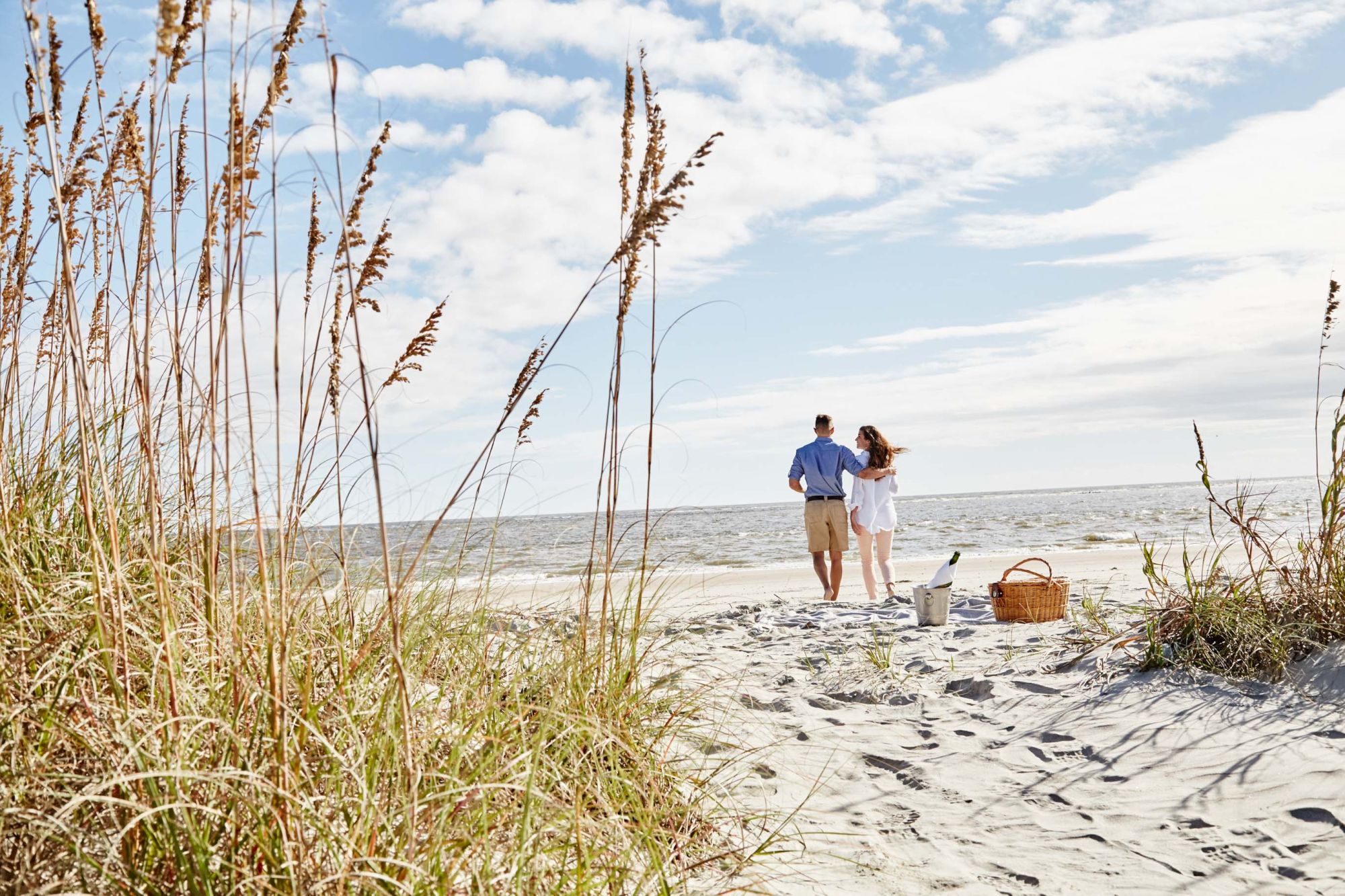 A couple walks along a sandy beach with ocean waves in the background, beside a picnic basket and a bottle on the shore.