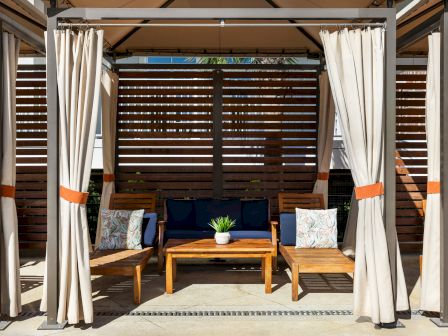 A cozy outdoor seating area with curtains, wooden chairs, a table, and a blue couch, set against a slatted wooden backdrop.