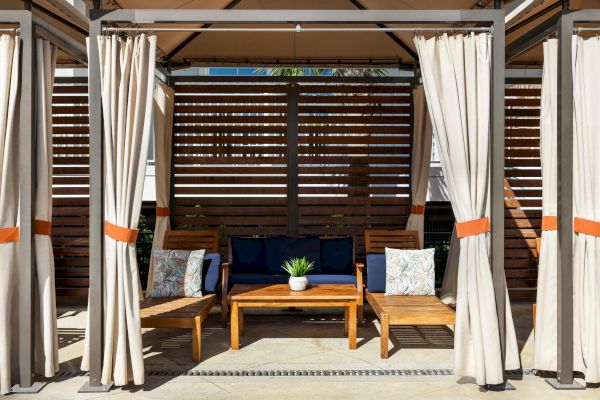 A cozy outdoor seating area with curtains, wooden chairs, a table, and a blue couch, set against a slatted wooden backdrop.