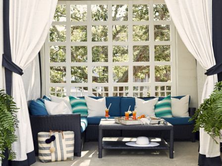 A cozy outdoor seating area with white curtains, blue and white pillows, a table with drinks, and lush ferns on both sides.