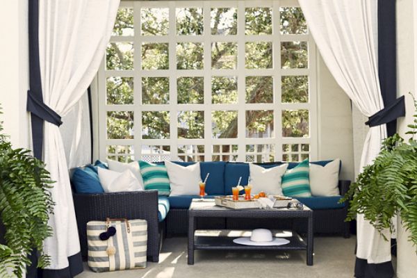 A cozy outdoor seating area with white curtains, blue and white pillows, a table with drinks, and lush ferns on both sides.