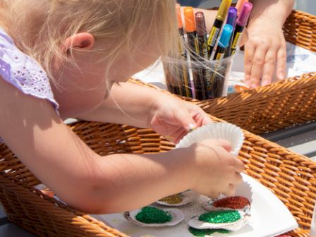 A child is decorating paper with glitter, using markers and paints, with art supplies in baskets.