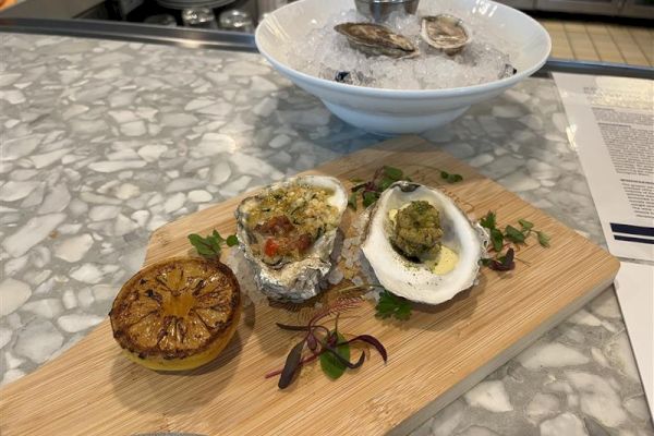 Grilled oysters with herbs and a charred lemon on a wooden board in front of a bowl with fresh oysters on ice.