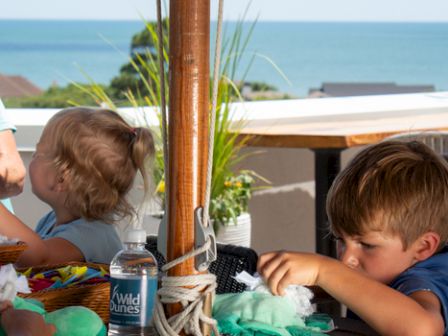 A woman is with two children engaging in crafts on a terrace with a sea view, featuring materials, a water bottle, and sunlit seating.