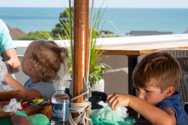 A woman is with two children engaging in crafts on a terrace with a sea view, featuring materials, a water bottle, and sunlit seating.