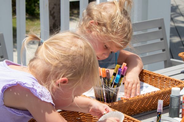 Two children are engaged in a craft activity with markers and colored items in wicker baskets on an outdoor table.