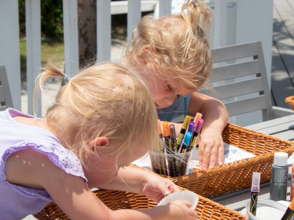 Two children are engaged in a craft activity with markers and colored items in wicker baskets on an outdoor table.
