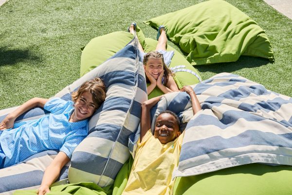 Three kids are lying on large green and striped cushions outdoors, smiling and relaxing on a grassy surface in the sunshine.