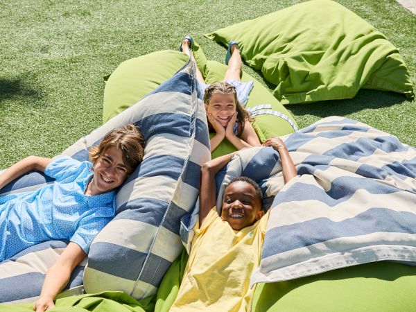 Three kids are lying on large green and striped cushions outdoors, smiling and relaxing on a grassy surface in the sunshine.