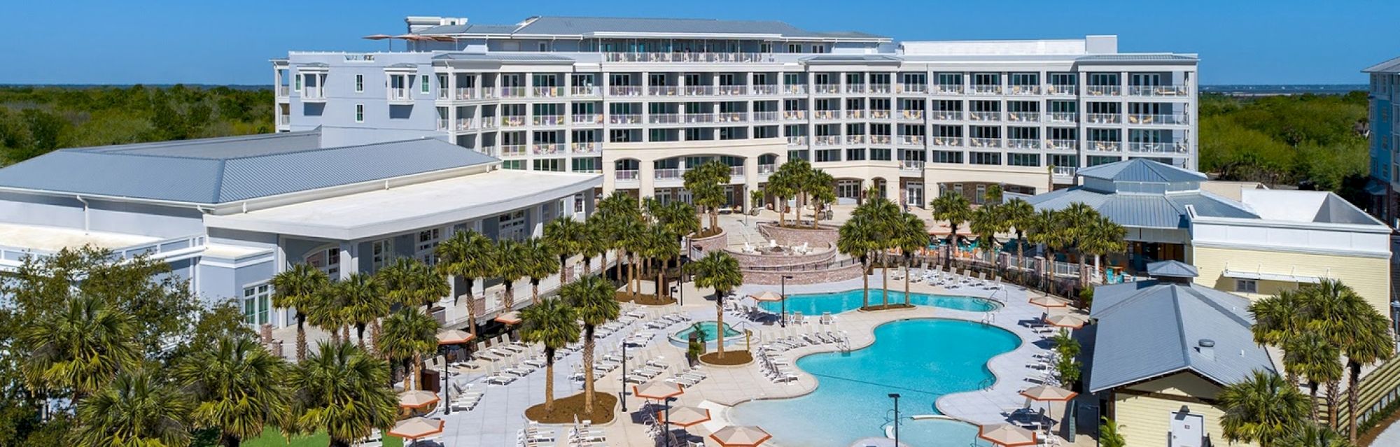 A tropical resort features palm trees, a pool with lounge chairs, and a large, multi-story building under a clear blue sky.