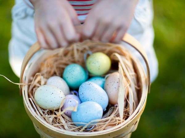 A person holding a basket with pastel-colored eggs on a bed of straw, suggesting a festive or Easter theme.