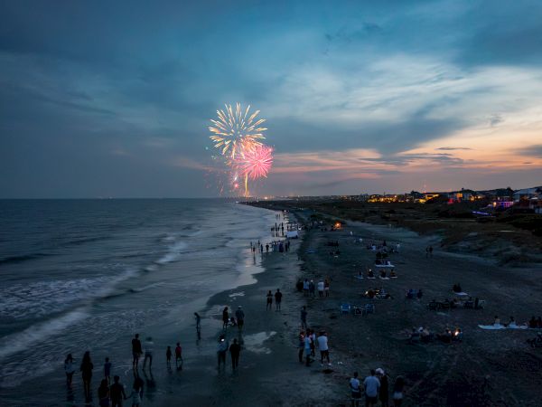 A beach at dusk with people gathered along the shore, watching colorful fireworks in the sky.