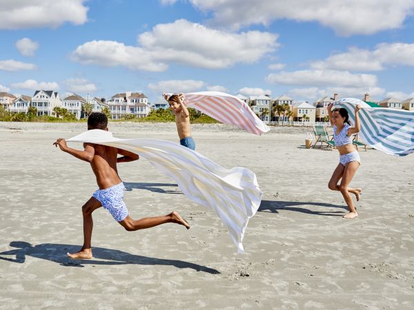 Children are running on the beach with towels in the air, under a partly cloudy sky, with houses in the background.