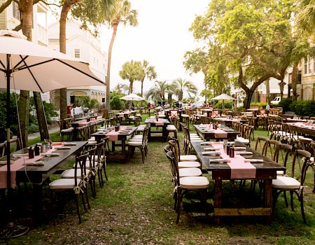 The image shows an outdoor dining setup with long tables, chairs, umbrellas, and pink table runners on a grassy area between buildings.